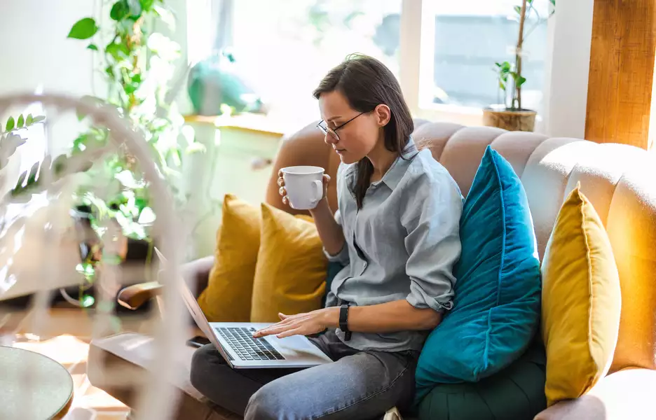 Une femme sur son canapé qui boit dans une tasse et travaille sur son ordinateur portable